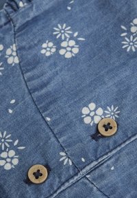 Tissu en denim avec un fond bleu présentant des motifs floraux blancs. Deux boutons ronds en bois sont visibles, avec une surface texturée et quatre trous.