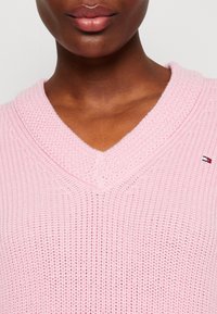 Maglione lavorato a maglia rosa con scollo a V. Presenta un tessuto testurizzato con un piccolo logo sulla parte sinistra.