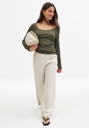 Långärmad olivgrön ribbad topp, krämfärgade vida byxor, svarta flip-flops, håller i en beige ficka. Slät textur, avslappnad passform.