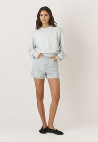 Šviesiai pilkas trumpas sweatshirt'as su raukšlėtais rankogaliais, derinamas su džinsiniais šortais. Modelis dėvi juodus šlepetės tipo batus. Paprasta, kasdienė apranga.