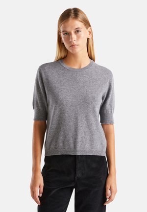 Junge Frau mit glatten blonden Haaren trägt einen grau kurzärmeligen Pullover und schwarze Hosen, steht vor einem schlichten weißen Hintergrund.