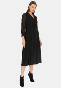Czarna sukienka z dopasowaną talią i plisowanym dołem. Posiada przezroczyste długie rękawy i dekolt w kształcie litery V. Stylizowana z czarnymi botkami za kostkę.