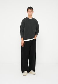 Dunkelgrauer Pullover mit rundem Ausschnitt, über einem weißen Hemd getragen, kombiniert mit lockeren schwarzen Hosen und beigen Turnschuhen.