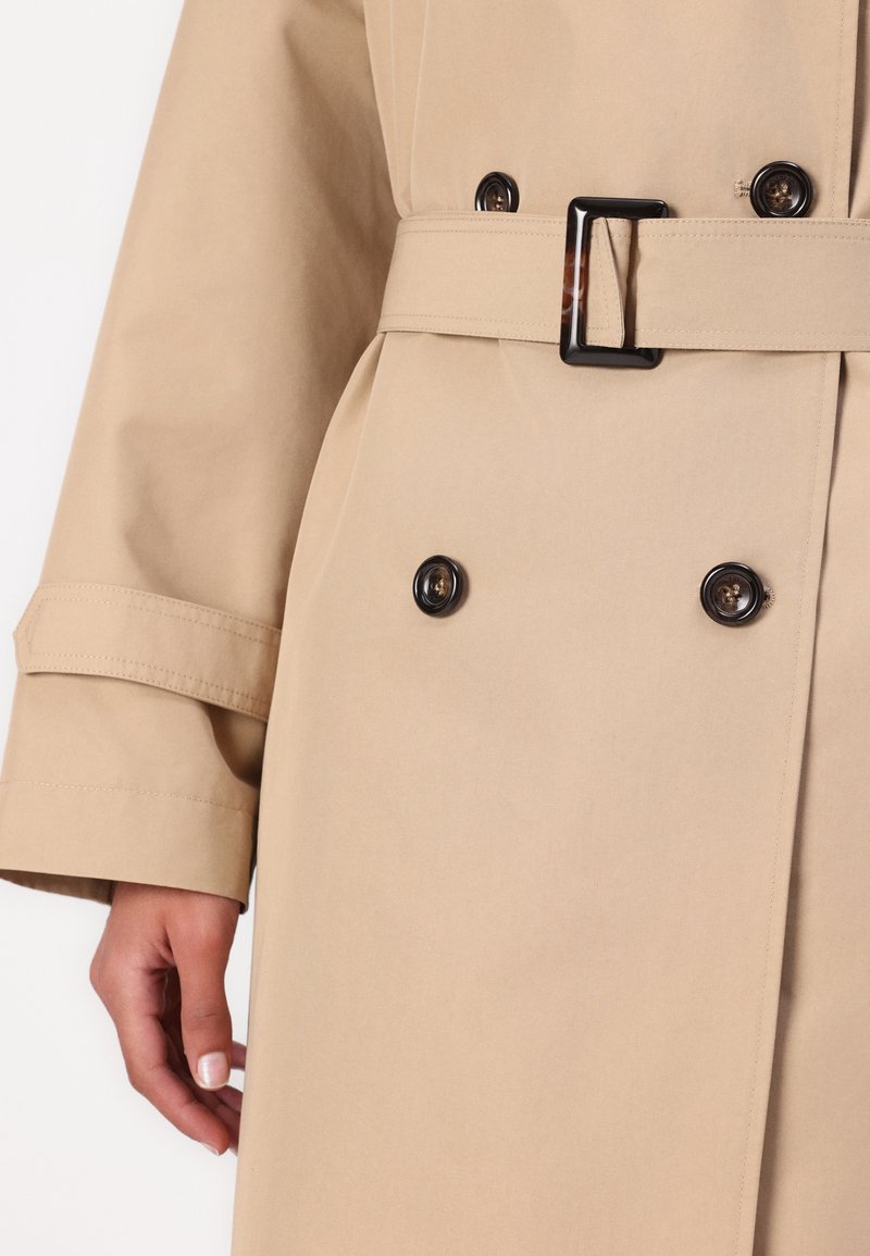 Beige Trenchcoat mit tailliertem Gürtel, ausgestattet mit schwarzen Knöpfen und einem glatten, strukturierten Stoff. Die Bündchen haben eine maßgeschneiderte Optik mit Nähdetails.