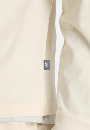 Sweat à la couleur crème avec une texture lisse, présentant un ourlet arrondi et une petite étiquette noire sur le côté.