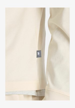 Cremfarbener Pullover mit glatter Textur, abgerundetem Saum und einem kleinen schwarzen Logo-Tag an der Seite.
