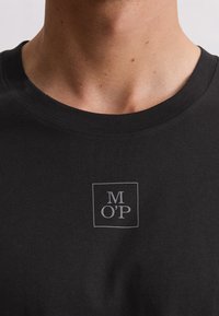 Close-up van een persoon die een zwart t-shirt draagt met een klein grijs "M O' P" logo in een vierkant op de borst.