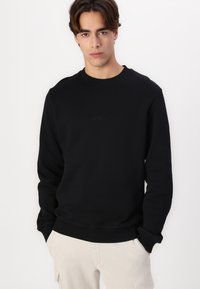 Zwarte sweatshirt van zacht materiaal, met een ronde hals, lange mouwen, ribgebreide manchetten en zoom, en subtiele reliëf logo-detail aan de voorkant.