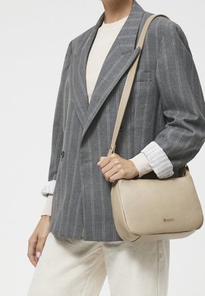 Personne portant un blazer gris à rayures fines et un pantalon blanc, tenant un sac à bandoulière en cuir beige avec une sangle courte sur un fond blanc.