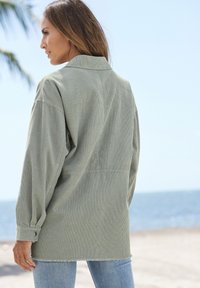 Overmaatse olijfgroene shirt met een corduroytextuur, knoopmanchetten en een rafelige zoom, gecombineerd met lichtblauwe jeans.