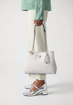 Borsa bianca a trama con manici a catena argentata, logo frontale e un piccolo cartellino, abbinata a scarpe con tacco argentate e pantaloni color crema.
