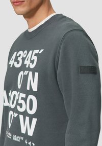 Szary sweatshirt z wyraźnym białym tekstem z współrzędnymi geograficznymi. Tekst jest wyraźnie widoczny na klatce piersiowej, miękka tkanina i rypsowany kołnierz.
