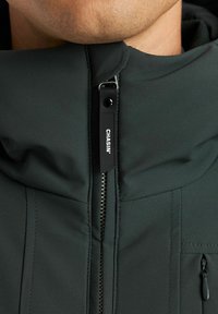 Chaqueta verde oscura con un alto cuello, cierre de cremallera y un tirador de cremallera con marca. Presenta una tela suave y un pequeño bolsillo con cremallera en el lado izquierdo.