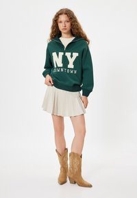 Sudadera verde con letras blancas "NY DOWNTOWN", combinada con una falda plisada beige y botas de cowboy marrones con costuras decorativas.