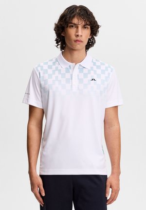 Młody mężczyzna o falujących ciemnych włosach, ubrany w biały poli z jasnoniebieskim wzorem w kratę na klatce piersiowej.