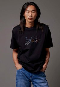 Zwarte katoenen T-shirt met kleurrijke grafische prints van vogels en sterren. Model draagt blauwe jeans en staat met de handen in de zakken.