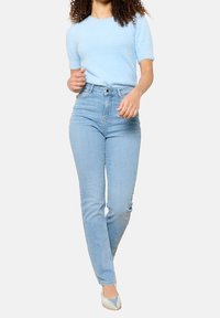 Světle modrý chlupatý svetr s krátkým rukávem v kombinaci s vysokým pasem světle modrých džín, které mají rovný střih a minimální ozdoby.