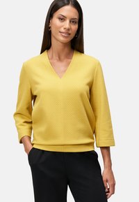 Blouse jaune texturée avec un col en V et des manches trois-quarts, associée à un pantalon noir. Le tissu arbore un motif géométrique.