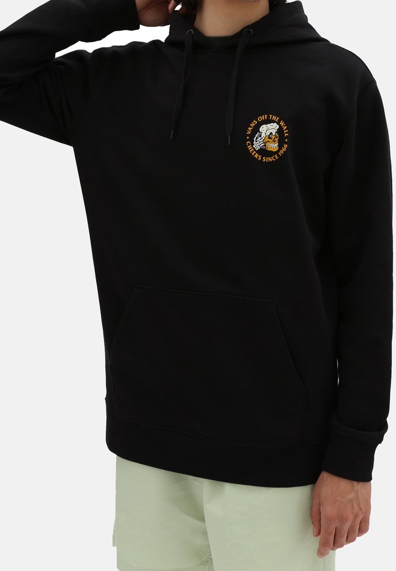 Personne portant un sweat à capuche noir avec une poche avant et un logo tête de mort orange "Vans Off The Wall Cheers Since 1966" sur la poitrine.