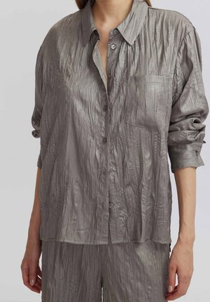 Osoba ubrana w luźną, pomarszczoną szarą koszulę na guziki z kieszenią na piersi oraz dołączone spodnie, z podwiniętymi rękawami do przedramion.