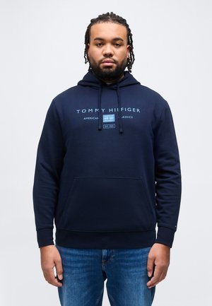 Man met gevlochten haar, gekleed in een marineblauwe Tommy Hilfiger hoodie en blauwe jeans, staand tegen een effen lichte achtergrond.