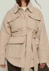 Beige uldblanding jakke med lange ærmer, store forlommer og et bundet bælte i taljen. Har en knaplukning foran og krave.