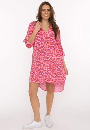 Woman smiling, wearing a pink and orange leopard print dress with rolled sleeves and white sneakers, standing against a white background.