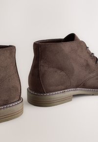 Hnedé členkové topánky z brúseného semišu so zaoblenou špičkou, sivými šnúrkami a textúrovanou gumovou podrážkou. Majú kontrastné prešívanie a polstrovaný opätok.