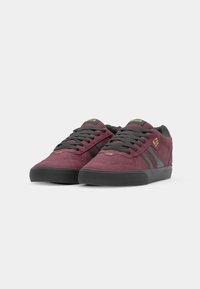 Burgundy suede sneakers with gray accents, featuring a round toe, flat rubber sole, and black laces. Gold logo on the tongue.