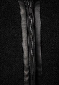 Svart jacka med dragkedja som har en mjuk, texturerad ullkropp och eleganta läderdetaljer längs dragkedjan. Högkontrastmaterialen smälter samman sömlöst.