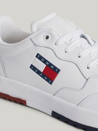 Sneakers brancos feitos de pele sintética, apresentando um patch de logo colorido com "TOMMY JEANS". Sola texturizada com detalhes em vermelho e azul-marinho.