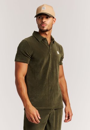 Olivgrünes Poloshirt mit weicher Textur, zwei Frontknöpfen und einem Logo-Patch. Kombiniert mit einer beigen Kappe mit Logo.