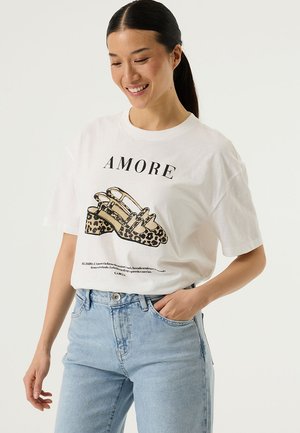 Wit katoenen T-shirt met een zwarte afbeelding van schoenen met luipaardprint en het woord "AMORE" in opvallende letters erboven. Losse pasvorm, korte mouwen.