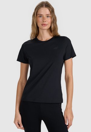 Młoda kobieta z długimi falującymi włosami, ubrana w prosty czarny t-shirt z krótkim rękawem i czarne spodnie, stojąca na jasnoszarym tle.