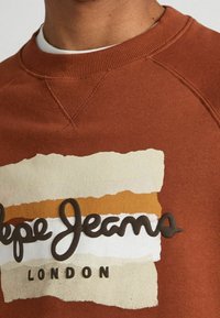 Primo piano di una felpa marrone con strati di pennellate beige, bianche e senape, e un logo testurizzato "Pepe Jeans London" sul petto.