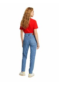Rotes Kurzarm-T-Shirt kombiniert mit hoch taillierten, hellblauen Jeans. Die Jeans verfügen über zwei Gesäßtaschen und haben leichte Abriebspuren, getragen mit weißen Turnschuhen.