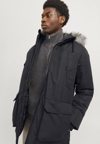 Parka noire avec capuche en fourrure, dotée d'une fermeture éclair et de boutons, de plusieurs poches à l'avant et d'un pull gris texturé en dessous.