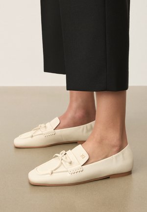 Pantofi loafers din piele crem cu detaliu fundiță, purtați cu pantaloni negri scurtați pe un podea de culoare bej deschis.