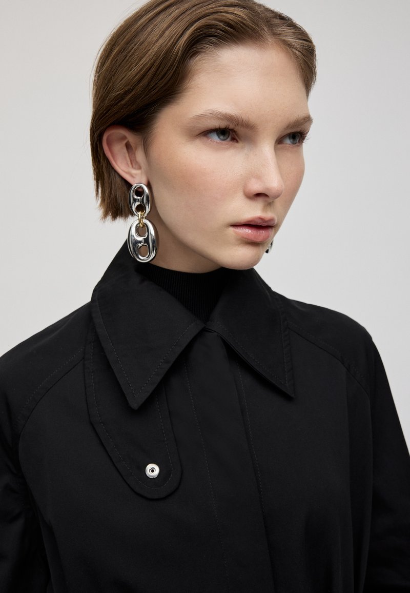 Schwarzer Mantel mit Kragen und Knopfdetails, kombiniert mit großen silbernen Ohrringen in Form von Schlaufen, die goldene Akzente und eine offene Mitte aufweisen.