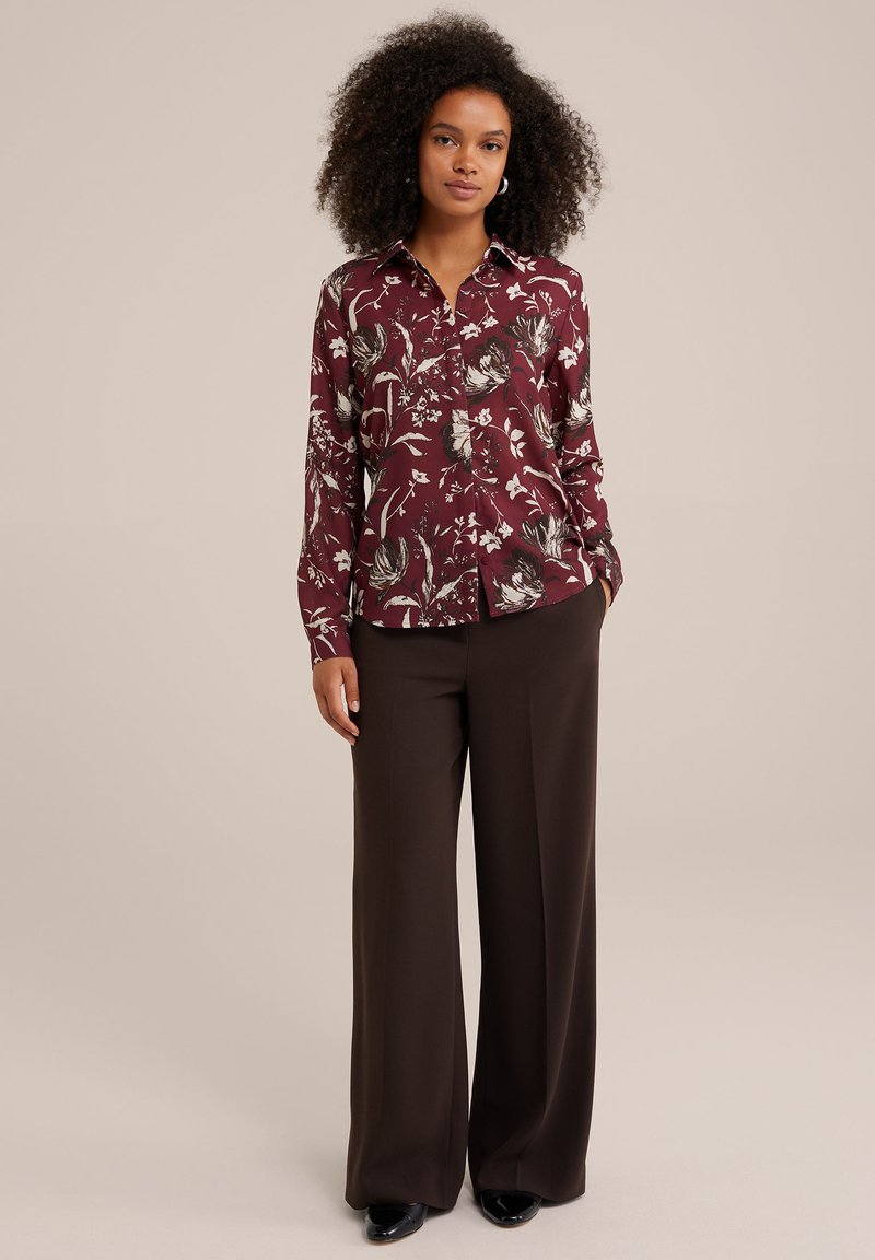 Burgundowa bluzka w kwiaty z długimi rękawami, z przodu zapinana na guziki, z kołnierzykiem, w połączeniu z brązowymi spodniami o szerokich nogawkach i czarnymi butami.