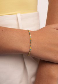 Dünnes, mehrfarbiges Perlenarmband mit türkisfarbenen, gelben und weißen Perlen, auf einem zarten Draht in Goldfarbe. Am Handgelenk getragen.