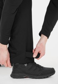 Pantalons légers noirs avec des sangles Velcro aux chevilles, associés à une chaussure de randonnée noire avec des côtés texturés et une semelle robuste.