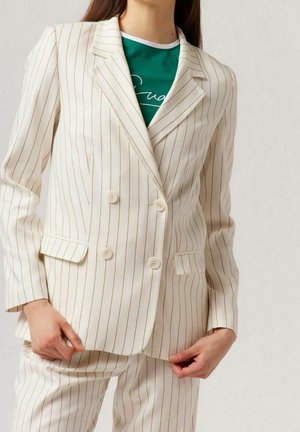 Woman wearing a cream suit with thin brown vertical stripes and a green top underneath, holding the jacket front with both hands.