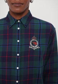 Chemise boutonnée à carreaux verts et bleu marine avec des accents rouges et bleus, présentant un design de blason orné de bijoux sur la poitrine gauche. Matériau en coton doux.