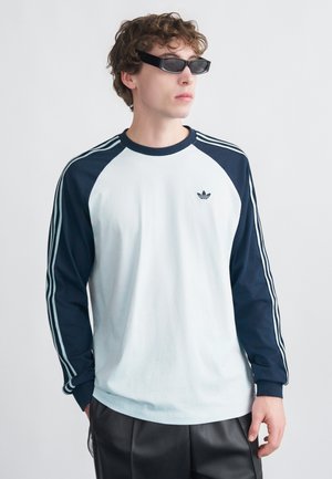 Lichtblauw T-shirt met lange mouwen en donkerblauwe raglanmouwen, met drie strepen op elke mouw en een klein logo op de borst.