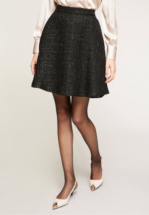 Kobieta w teksturowanej czarnej spódnicy w kształcie litery A, przezroczystych czarnych rajstopach, białych szpilkach z złotymi zakończeniami i jasnoróżowej jedwabnej bluzce.