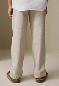 Persona con pantalones de lino beige y sandalias marrones, de pie sobre un suelo claro frente a un fondo neutro.