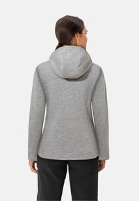 Vrouw in een aansluitende grijze hoodie en zwarte broek, gezien vanaf de achterkant tegen een effen achtergrond.
