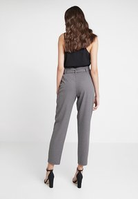 Grijze hoge taillebroek met een taps toelopend been, voorzien van een gladde textuur, riemlussen en een zwarte tanktop. Zwarte sandaaltjes met hak maken de look af.