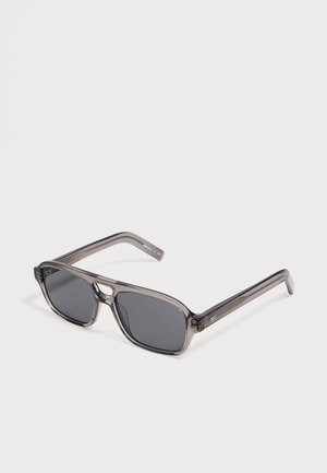 Lunettes de soleil rectangulaires gris foncé avec monture en plastique gris translucide et verres noirs posées sur une surface blanche.
